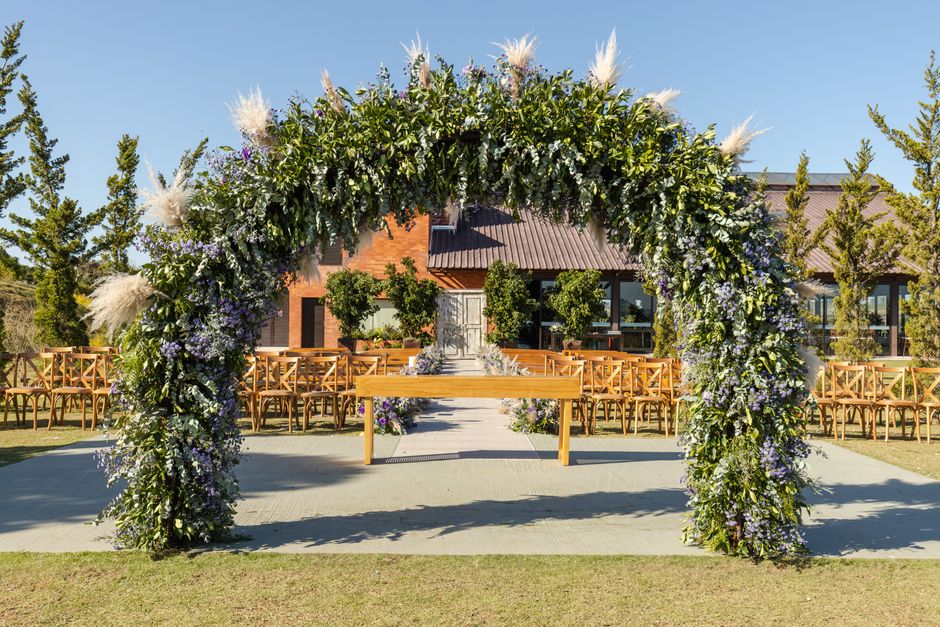 A large floral arch decorated with a lush arrangement of white pampas grass, purple and white flowers, and green foliage, positioned outdoors on a paved area in front of a brick building with glass wi